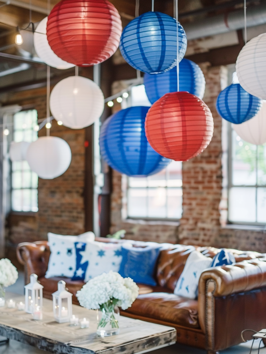 Fourth of July themed loft party. Sunlit loft space with hanging red, white, and blue paper lanterns, a distressed leather couch arrayed with plush star-patterned throw pillows, and a rustic wood table set with mason jars filled with white hydrangeas and blue-dyed baby's breath.