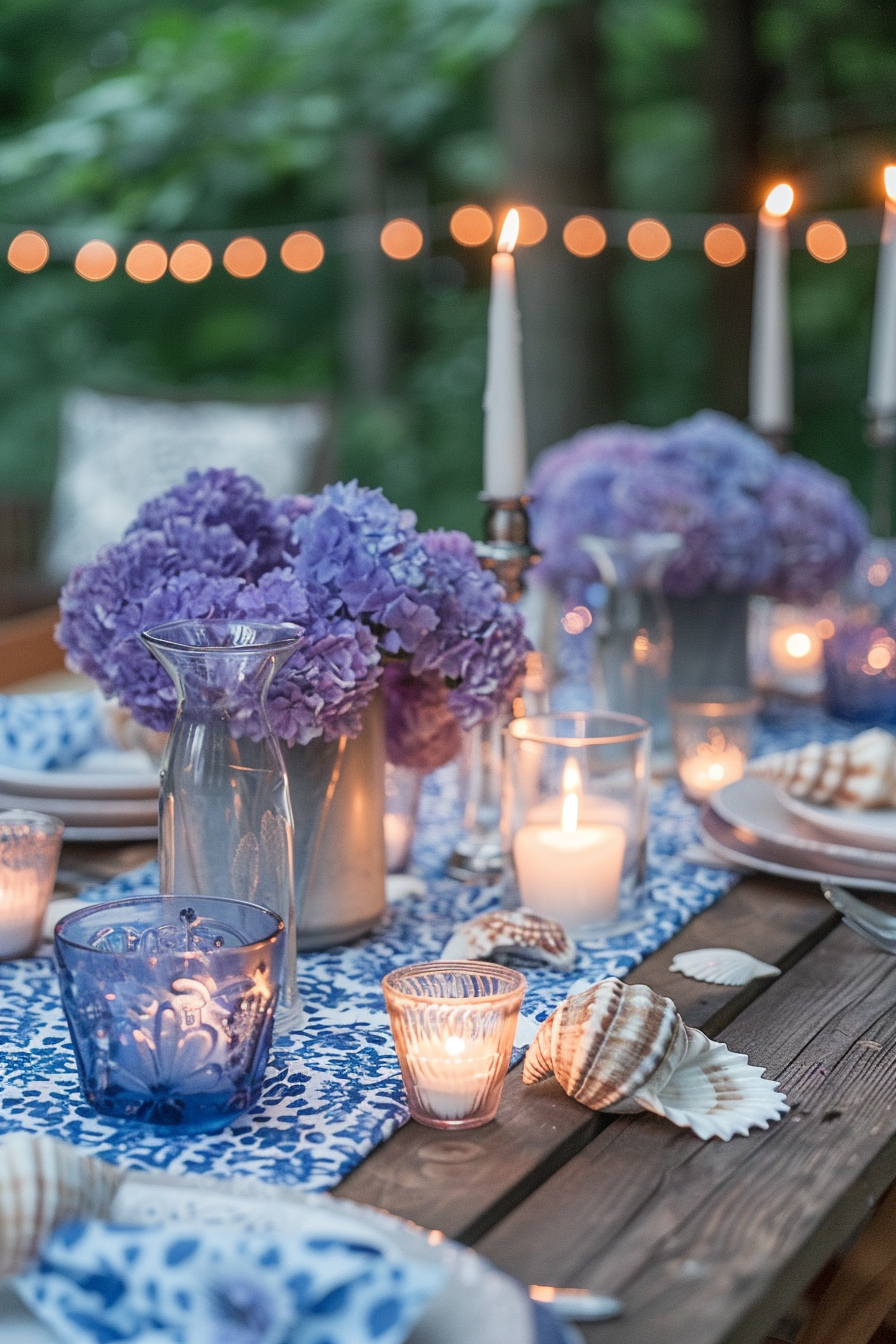 Backyard dinner party table decor. A rustic wooden table adorned with indigo and white block print tablecloth, oversized transparent hurricane candle holders, fresh purple hydrangea flower centerpiece, and handmade seashell place cards.