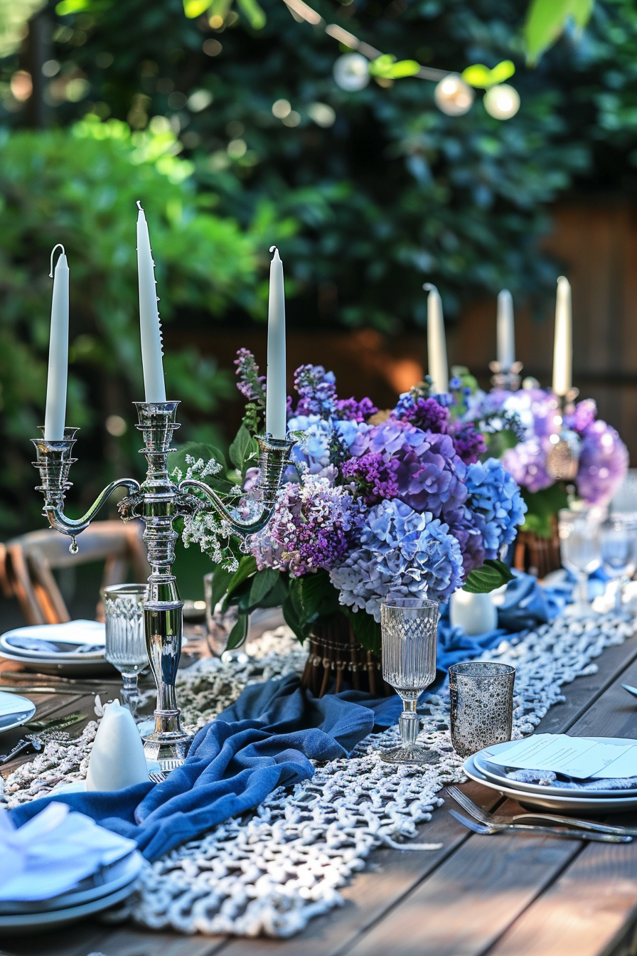 Backyard dinner party table decor. Navy blue macrame table runner, sleek silver candelabras, and freshly picked blend of lilac and hydrangea centerpiece, arranged on a rustic wooden table.