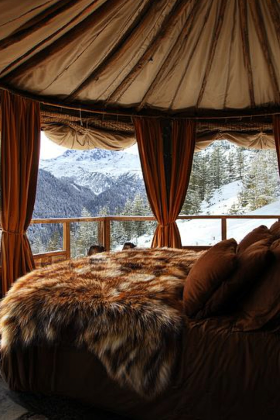 Alpine-view yurt bedroom. Rustic bed, panoramic windows and brown fur throws.